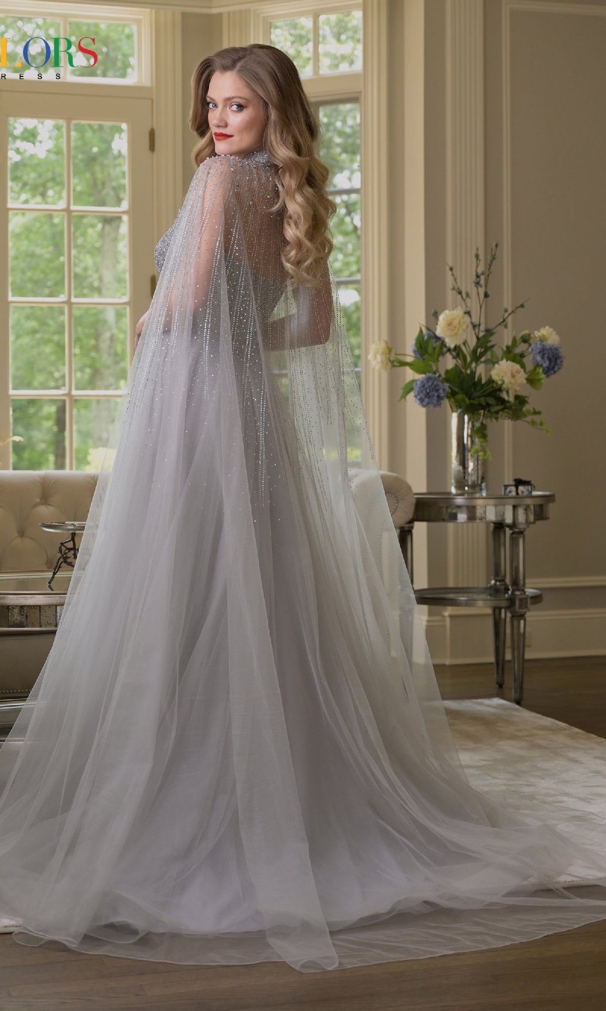 A woman stands indoors by a window, looking over her shoulder in the Colors Dress 3879—a light gray, floor-length prom dress with an embellished corset bodice and sheer beaded cape.